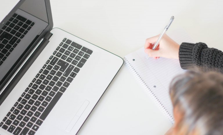 person in front of a laptop screen writing on loose leaf paper