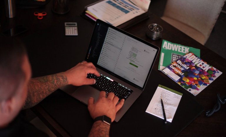 desk with issues of Adweek, a notepad, and a computer showing the overhead marketing tasks