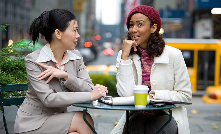 two women sitting at a table wearing business attire outside in a city environment with a cup of coffee and notebook