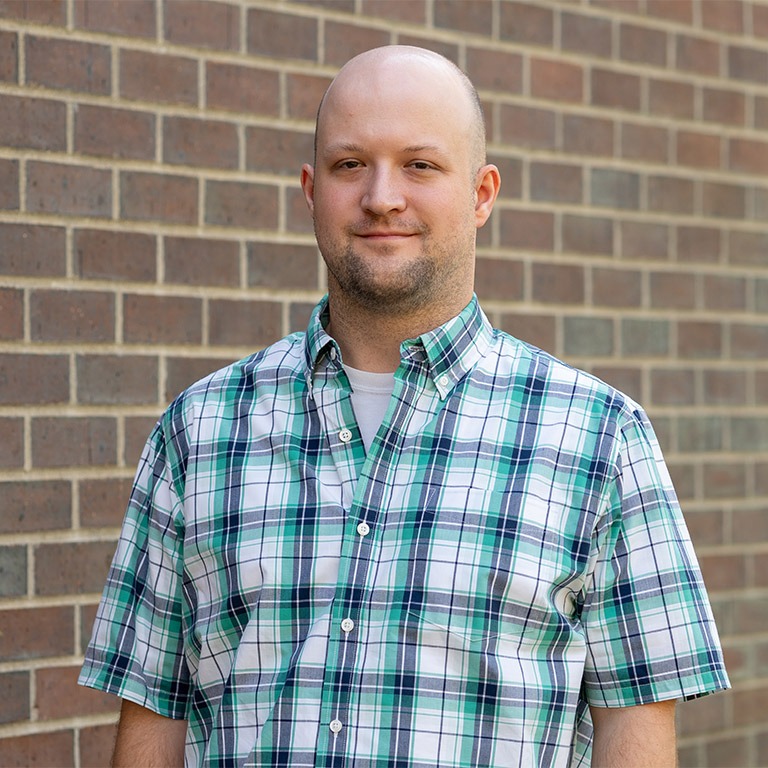 adult male wearing a blue and white plaid shirt standing in front of a brick wall