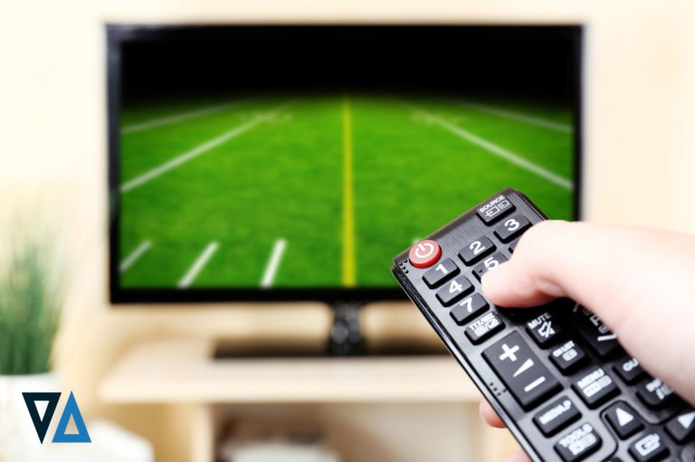 adult hand holding a black remote pointed at a black television with a football field on the screen