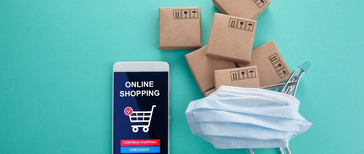 white iPhone with a grocery shopping app opened on the screen next to a miniature shopping cart filled with brown packages and covered with a blue face mask