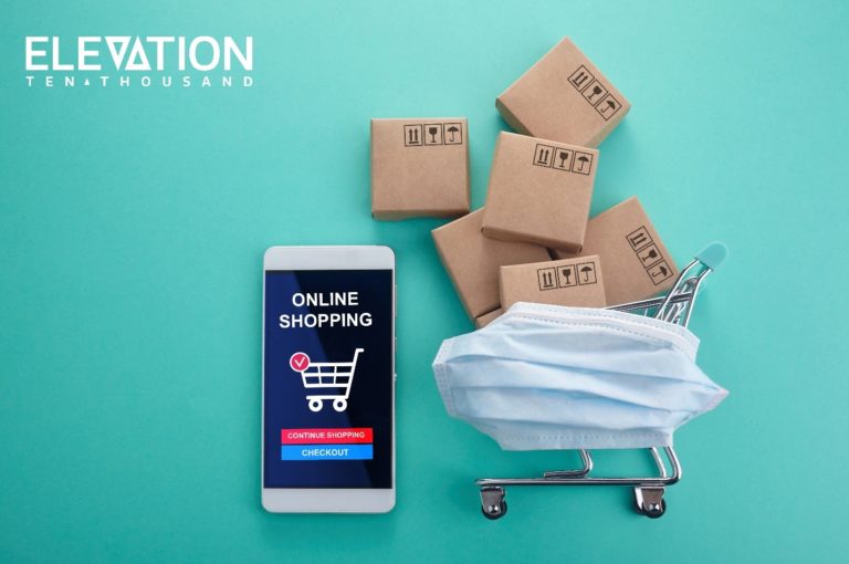 white iPhone with a grocery shopping app opened on the screen next to a miniature shopping cart filled with brown packages and covered with a blue face mask