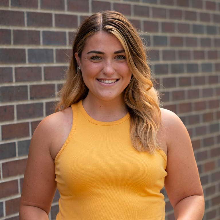 blonde woman in yellow top standing in front of brick wall