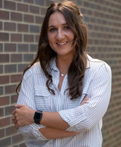 woman with long brown hair and white plaid button-down shirt standing in front of brick wall