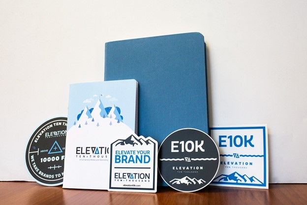 blue notebook in front of a white wall with Elevation Ten Thousand-branded coaster, notebook, stickers, and magnet
