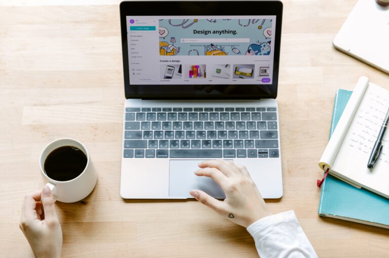 overhead shot of person at light brown, wooden desk holding white mug of black coffee designing a website on a silver laptop