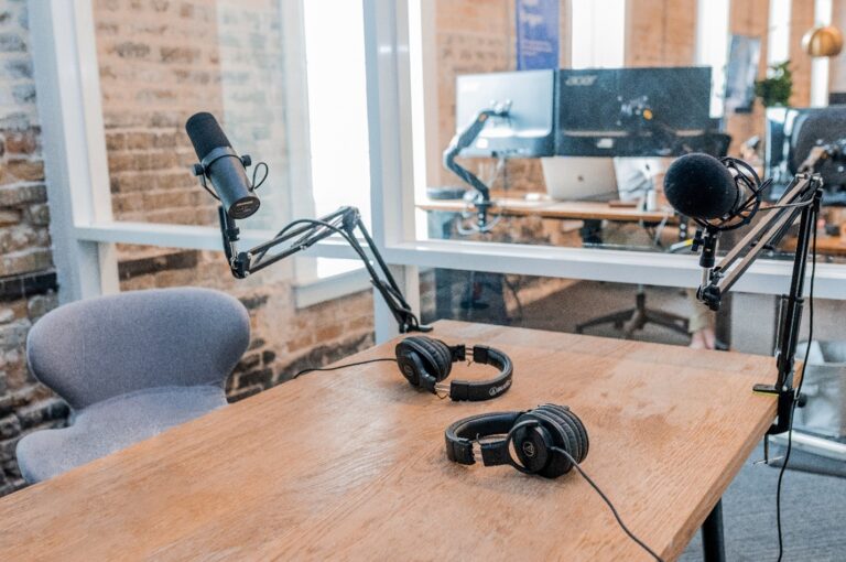 wooden desk with a gray desk chair, black headphones, and black microphones used to record a podcast