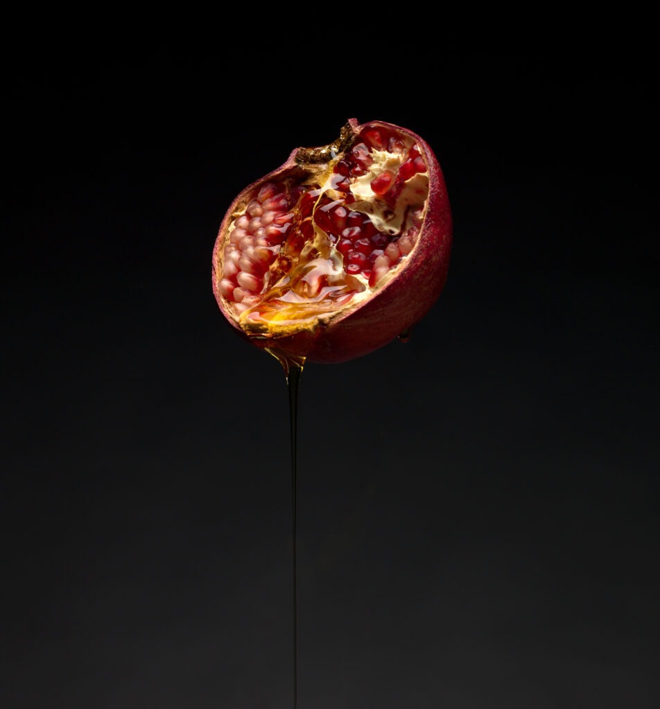 red pomegranate cut in half showing red seeds with honey dripping over it on a black background