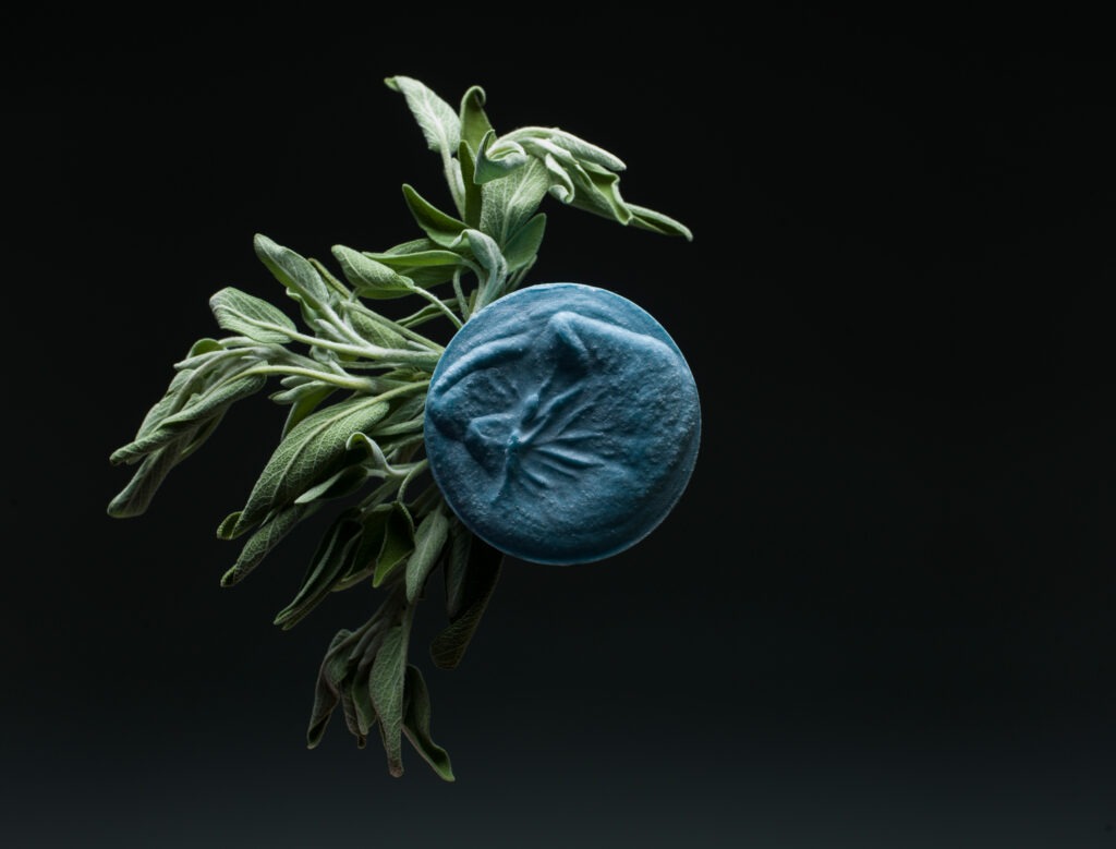 green sage leaves next to a round, blue bar of avitals soap on a black background