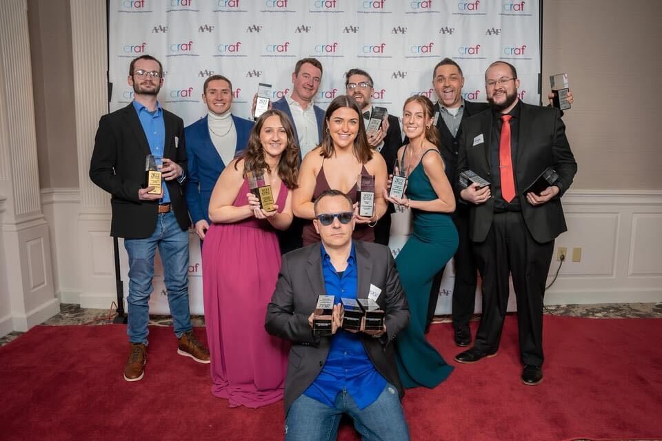 group of 10 adults in dress clothes holding trophies on a red carpet