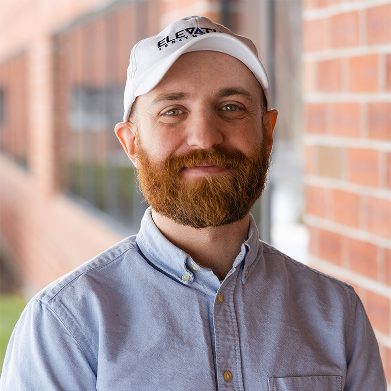 man wearing a blue button up shirt and white baseball hat by a brick building